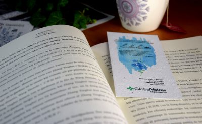 Sur un bureau, un livre ouvert où est posé le marque-page Global Voices esperanto. On y voit du texte sur fond d'encre bleue.