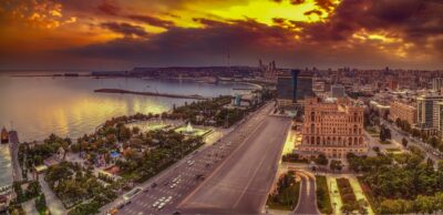 The image shows a part of Baku, capitol of Azerbaijan. The center of the image depicts a street that is lined on the one side by the ocean and the other by tall buildings and green parks. The upper part of the image shows the sky in a sunset setting with clouds. The further back of the image shows the city panorama.